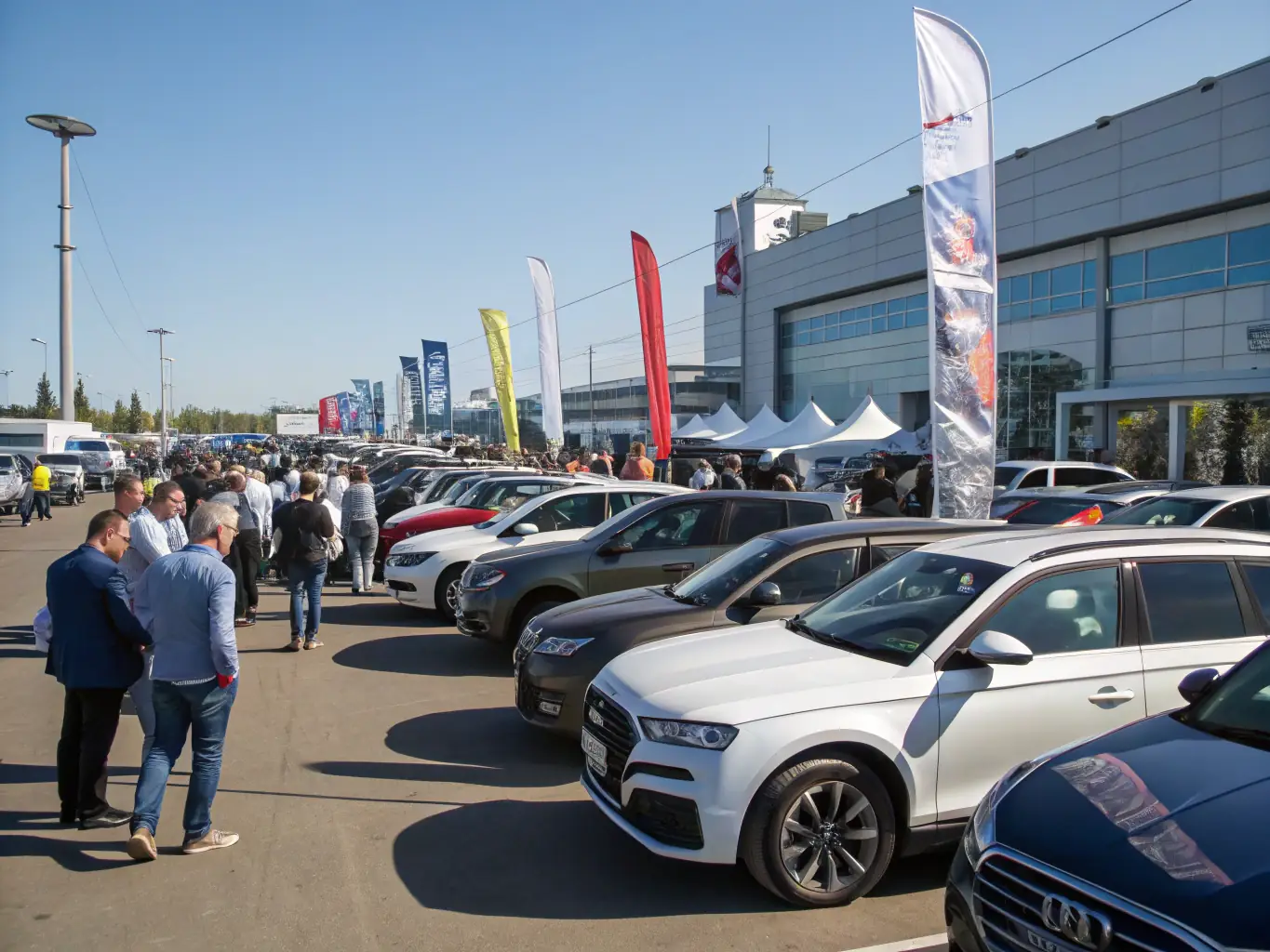A vibrant image of a bustling US car auction lot, filled with various makes and models of cars, under a clear blue sky. Banners and auction signs are visible in the background.